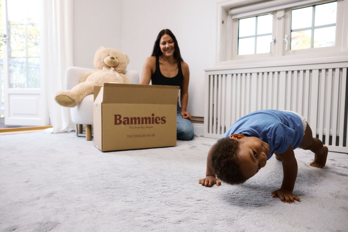 Glad mamma packar upp en kartong med Bammies blöjor i ett ljust barnrum medan ett busigt småbarn i blå t-shirt och blöja tittar fram mellan benen på golvet i förgrunden.