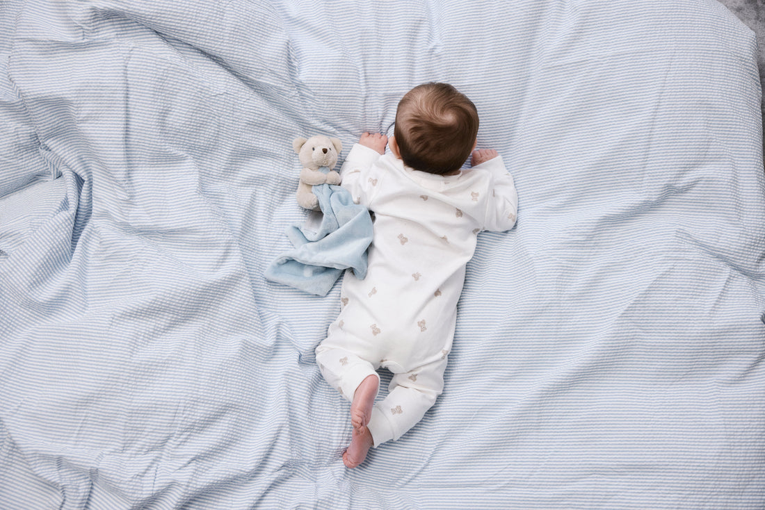 Bebis i vit pyjamas med nallemönster ligger på mage på ett blåvitrandigt påslakan, bredvid en snuttefilt med nalle, fotograferat ovanifrån.