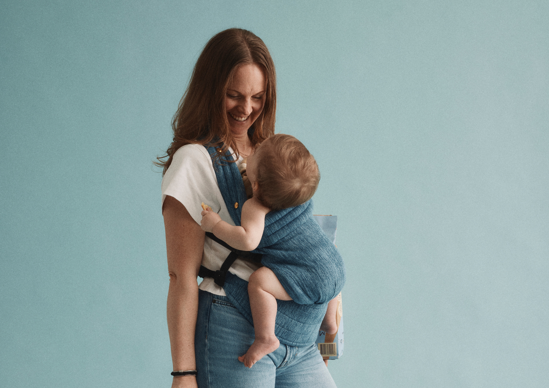Förälder bär leende bebis med Bammies The Toddler i bärsele mot blå bakgrund 