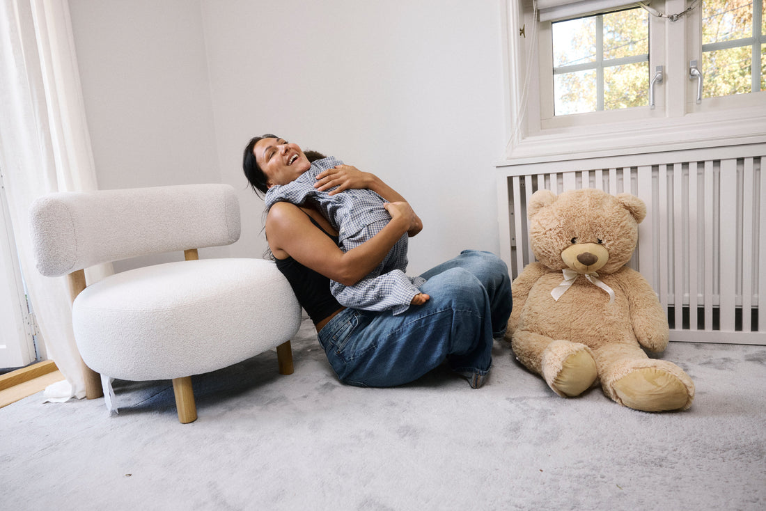 Glad mamma sitter på golvet i ett ljust barnrum och kramar sitt barn i rutig babydräkt, bredvid en vit fåtölj och en stor nallebjörn